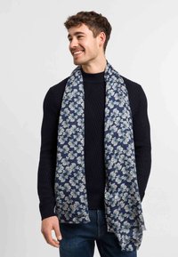 Smilende mand iført en sort ribbet sweater og blå jeans, draperet med et marineblåt tørklæde med et hvidt blomsterdesign.