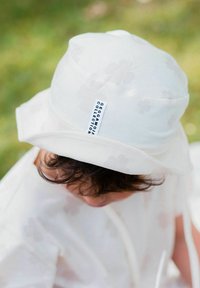 Lapsi, jolla on kiharat hiukset, käyttää valkoista ämpärihattua, jossa lukee "Geggamoja Collection", ja siihen sopivaa vaaleata paitaa, ulkona nurmikolla.