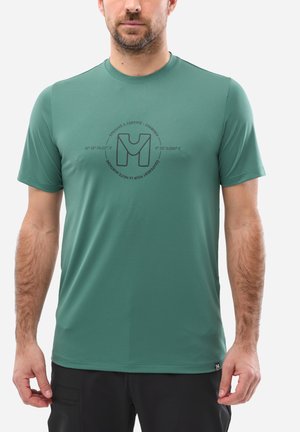 Homme portant une chemise verte à manches courtes avec un logo circulaire et du texte sur la poitrine, accompagné d'un pantalon noir, debout devant un fond blanc.