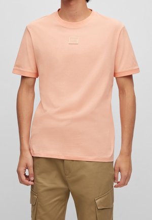 Person iført en ensfarvet ferskenfarvet T-shirt med korte ærmer med et lille "Hugo"-logo på brystet og khaki cargobukser.