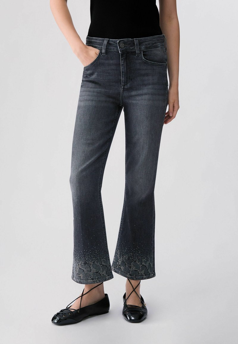Femme portant un haut noir ajusté, un jean évasé gris avec des détails en strass aux ourlets, et des ballerines noires à brides croisées.