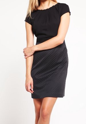 Femme portant une robe noire ajustée à manches courtes avec une jupe texturée à motifs, debout les bras croisés sur la taille.