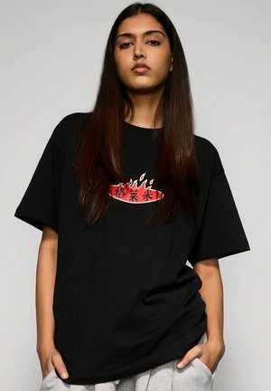 Młoda kobieta z długimi ciemnymi włosami, ubrana w czarny, oversize'owy T-shirt z czerwonym płomieniem i azjatyckimi znakami, z rękami w kieszeniach jasnoszarych spodni.