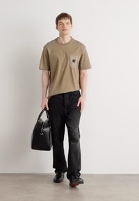 Tricou din bumbac bej cu un buzunar aplicat, asociat cu blugi negri. Modelul ține o geantă din piele neagră și poartă pantofi negri.