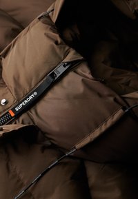 Brun pufferjakke-interiør med en glat tekstur, sort 'SUPERDRY'-mærke og kontrasterende syninger langs lynlåsen og sømmene.