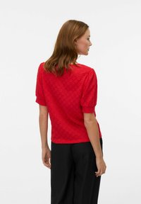 Blusa rossa con texture, maniche a palloncino corte e scollatura rotonda, abbinata a pantaloni neri, che mostrano un fit casual visto da dietro.
