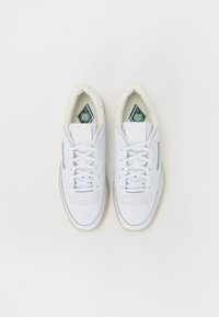 Λευκά αθλητικά αθλητικά παπούτσια από δέρμα με διάτρητες μύτες, επίπεδα κορδόνια και μαλακή επένδυση· περιλαμβάνει πράσινες λεπτομέρειες στο λογότυπο και τις εσωτερικές σόλες.