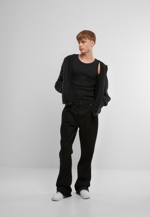 Un débardeur noir sous un sweat à capuche noir zippé, associé à un jean noir ample et des baskets blanches. Un design simple et monochromatique.