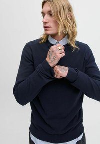 Suéter de un oscuro azul marino con cuello redondo y puños acanalados, usado sobre una camiseta de cuello con rayas; textura suave y diseño ajustado.