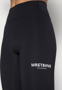 Černé sportovní legíny s vysokým pasem, hladkou texturou a bílým logem "WRSTBHVR ATHLEISURE" na dolní levé straně.