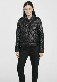 Czarna pikowana kurtka puchowa z okrągłym kołnierzem, błyszczącym wykończeniem, prążkowanymi mankietami i dopasowanym dołem. Nosi się ją na ciemne spodnie.