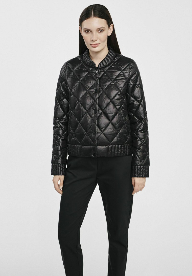 Czarna pikowana kurtka puchowa z okrągłym kołnierzem, błyszczącym wykończeniem, prążkowanymi mankietami i dopasowanym dołem. Nosi się ją na ciemne spodnie.