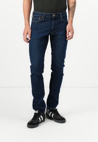 Jeans en denim bleu foncé avec une coupe slim, comportant cinq poches, des baskets noires et blanches, et un t-shirt uni de couleur foncée.