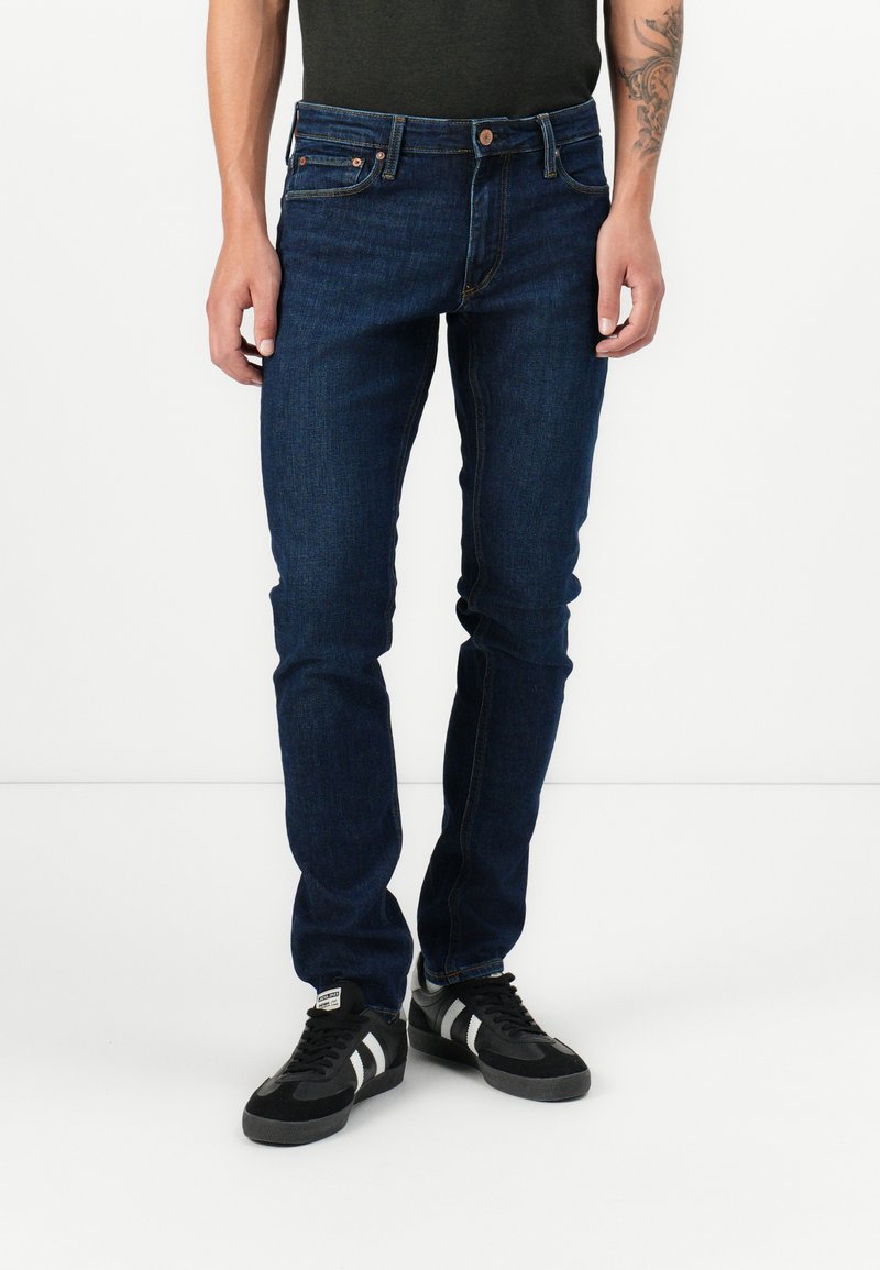 Jeans en denim bleu foncé avec une coupe slim, comportant cinq poches, des baskets noires et blanches, et un t-shirt uni de couleur foncée.