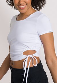 Crop top bianco a coste con maniche corte, caratterizzato da un cordino e aperture laterali. Accentuato da un'etichetta con logo sulla manica.