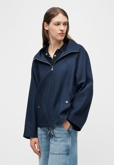 Γυναίκα που φοράει μπουφάν σε σκούρο μπλε χρώμα με φερμουάρ, ρυθμιζόμενο τελείωμα με κορδόνι και μπροστινές τσέπες, πάνω από μαύρο πουκάμισο με γιακά και ανοιχτόχρωμο μπλε τζιν.