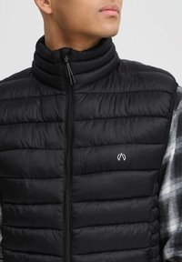 Gilet imbottito nero con colletto alto, texture trapuntata e zip frontale. Presenta un logo discreto sul petto. Indossato sopra una camicia a quadri.