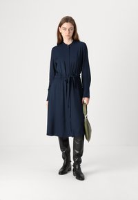 Marineblå, langermet kjole med knyt i midjen, knappet front og en avslappet passform. Kombinert med svarte knelengde støvler og en grønn handbag.
