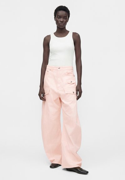 Γυναίκα που φοράει λευκό αμάνικο ριμπ τοπ, φαρδιά ανοιχτό ροζ παντελόνια cargo με καπάκια τσέπες και μαύρα παπούτσια με τετράγωνη μύτη, στέκεται μπροστά σε λευκό φόντο.