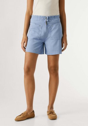 Shorts azul claro de talle alto con bolsillos frontales y cierre de doble botón, usados con camiseta sin mangas blanca y zapatos mocasines marrones.