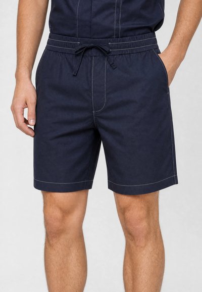 Homme portant un short bleu marine à cordon avec des coutures blanches, debout avec une main dans la poche devant un fond uni.