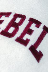 Hvitt stoff med fet burgunder brodert tekst som staver "REBELS." Teksturen er myk med synlige sømdetaljer.