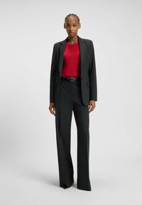 Costume nero a righe con giacca su misura e pantaloni a gamba larga, abbinato a un body rosso aderente. Tessuto liscio con una sottile texture.