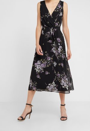Femme portant une robe midi noire sans manches avec un imprimé floral violet et blanc, col en V, ceinture à la taille, et des talons noirs à bride cheville.