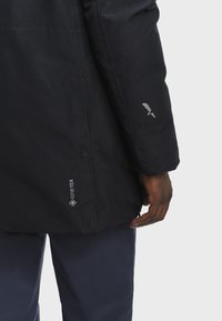 Close-up of a person wearing a black Gore-Tex jacket and dark pants, showing the lower sleeve and side of the jacket.