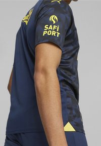 Musta urheilupaita, jossa on tekstuuripinta ja keltaisia korostuksia. Ominaisuuksiin kuuluu pyöreä kaula-aukko ja lyhyet hihat, ja paidassa on "SAFI PORT" -logo hihassa.