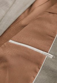 Interior de chaqueta de traje de mezcla de algodón con un forro de satén color beige con un patrón sutil, exterior a cuadros contrastantes y un bolsillo con cremallera.