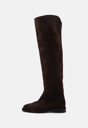 Une botte unique haute en daim marron foncé avec une semelle plate et un bout arrondi, vue de profil sur un fond uni.