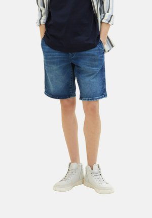 Jeanshorts in Dunkelblau, gerade geschnitten, mit umgeschlagenem Saum. Kombiniert mit weißen High-Top-Sneakern, aufrecht stehend mit hängenden Händen in den Taschen.