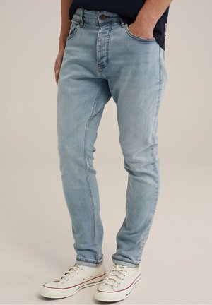 Homme portant un jean slim bleu clair avec des ourlets retroussés et des baskets en toile blanches, debout avec une main dans la poche.