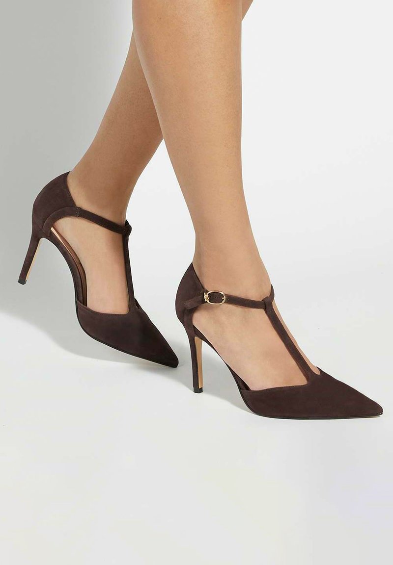 Escarpins en suede marron avec un bout pointu, un design à sangle en T et un talon aiguille mince, présentant un accent doré sur la sangle.