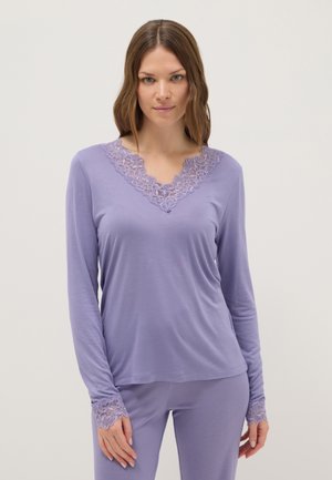 Donna che indossa una maglietta a maniche lunghe viola chiaro e pantaloni con rifinitura in pizzo sul collo a V e sulle maniche, in piedi davanti a uno sfondo chiaro e uniforme.