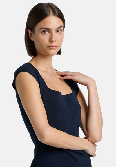 Mornarsko modra, prilagojena brezrokavna obleka s kvadratnim izrezom in mehko teksturo. Model pozira z eno roko na ramenu, z minimalističnim dizajnom.