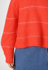 Punainen teksturoitu neulepaita pitkillä hihoilla, jossa on valkoisia vaakaraitoja, yhdistettynä siniseen denimihameeseen rennossa tyylissä.