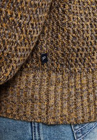 Chunky gebreide trui met bruine en gele garen, voorzien van een klein zwart etiket met een witte "P" detail, gecombineerd met blauwe spijkerbroek.