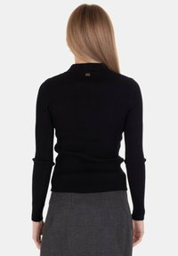 Donna con lunghi capelli biondi che indossa un top a maniche lunghe nero a coste e una gonna grigia, vista di spalle su uno sfondo neutro.