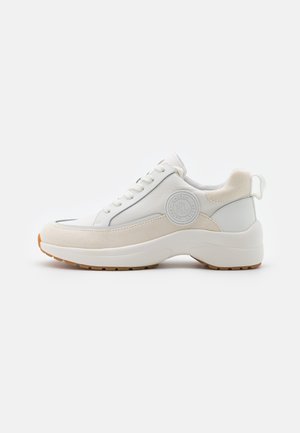 Baskets basses blanches et beiges avec semelle épaisse, lacets à l'avant et patch logo circulaire sur le côté, vues de l'extérieur.