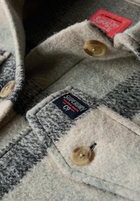 Wełniana koszula w szaro-kremową kratę, z zapięciem na guziki, okrągłym kołnierzykiem i metką Superdry. Miękka, teksturowana tkanina z widocznymi szwami.