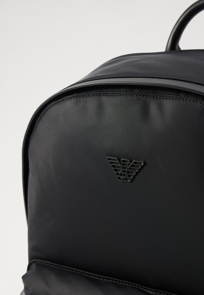 Mochila negra hecha de material sintético liso, con un emblema de logo brillante y una forma elegante y redondeada. Cierre con cremallera en la parte superior.