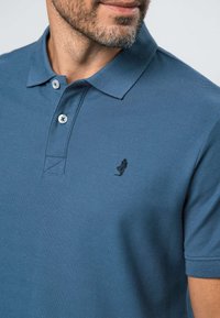 Parrakas mies, jolla on sininen lyhythihainen poolopaita, jossa on kaksi nappia ja pieni musta kirjailtu hahmo rinnassa.