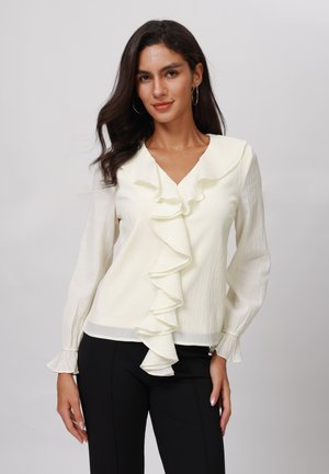 White ruffled blouse with a V-neck, long sleeves, and flared cuffs, made of textured fabric. Paired with black pants.