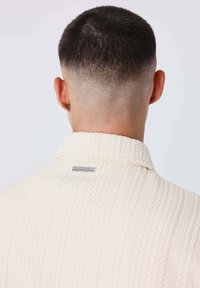 Cárdigan de punto texturizado en color crema, con cuello doblado, que presenta un patrón tejido y una etiqueta plateada de marca en la parte posterior del cuello.