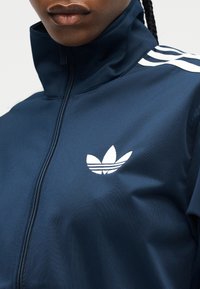 Nahaufnahme einer Person, die eine marineblaue Adidas-Zipjacke mit weißen Streifen auf den Schultern und dem Markenzeichen Trefoil auf der Brust trägt.