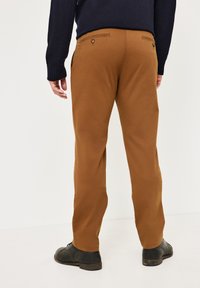 Braune Baumwollhose mit geradem Schnitt, ausgestattet mit zwei Gesäßtaschen mit Knöpfen und einer glatten Textur. Getragen mit schwarzen Schuhen.