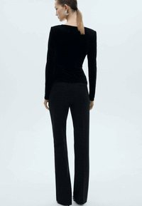 Zwarte fluwelen blouse met lange mouwen, gecombineerd met zwarte broeken. De blouse heeft een aansluitend ontwerp en een gladde textuur, wat een strakke silhouet creëert.