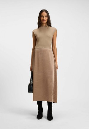 Strickkleid mit einem figurbetonten beigen Oberteil und einem schwingenden beigefarbenen Satinrock, kombiniert mit schwarzen High-Heels und einer schwarzen Handtasche.